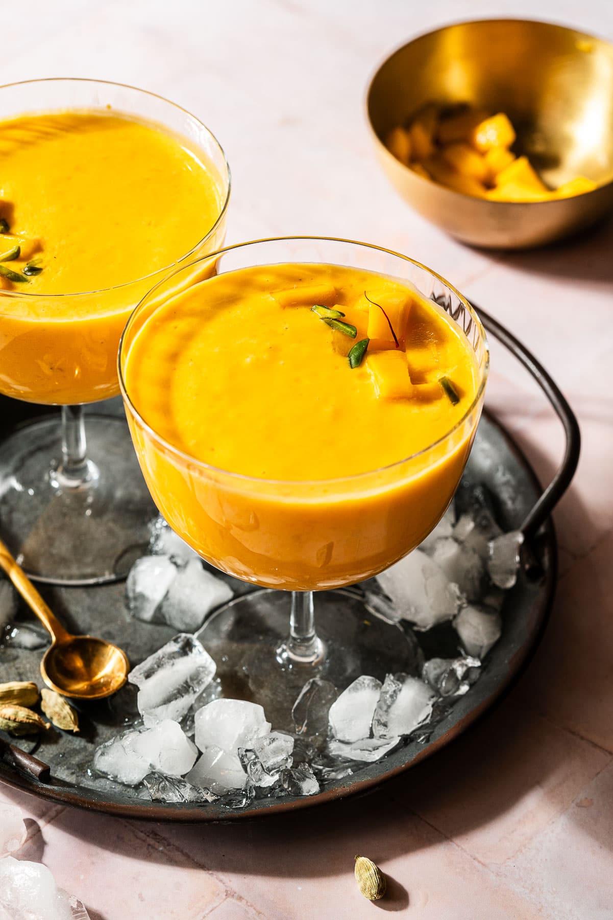 Two glasses of mango lassis in a platter with green cardamom pods.
