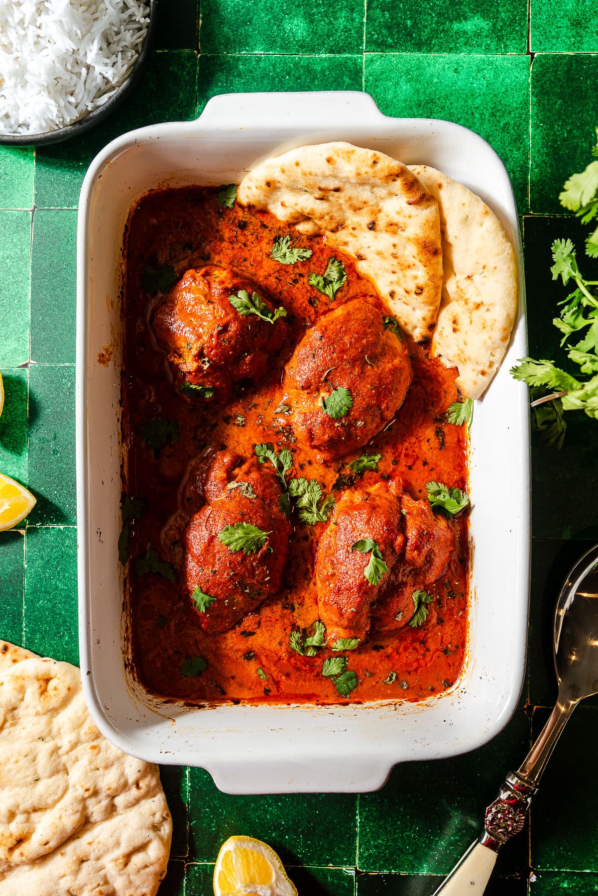 A Baking dish filled with four chicken thighs in a butter chicken sauce with naan and rice for serving.