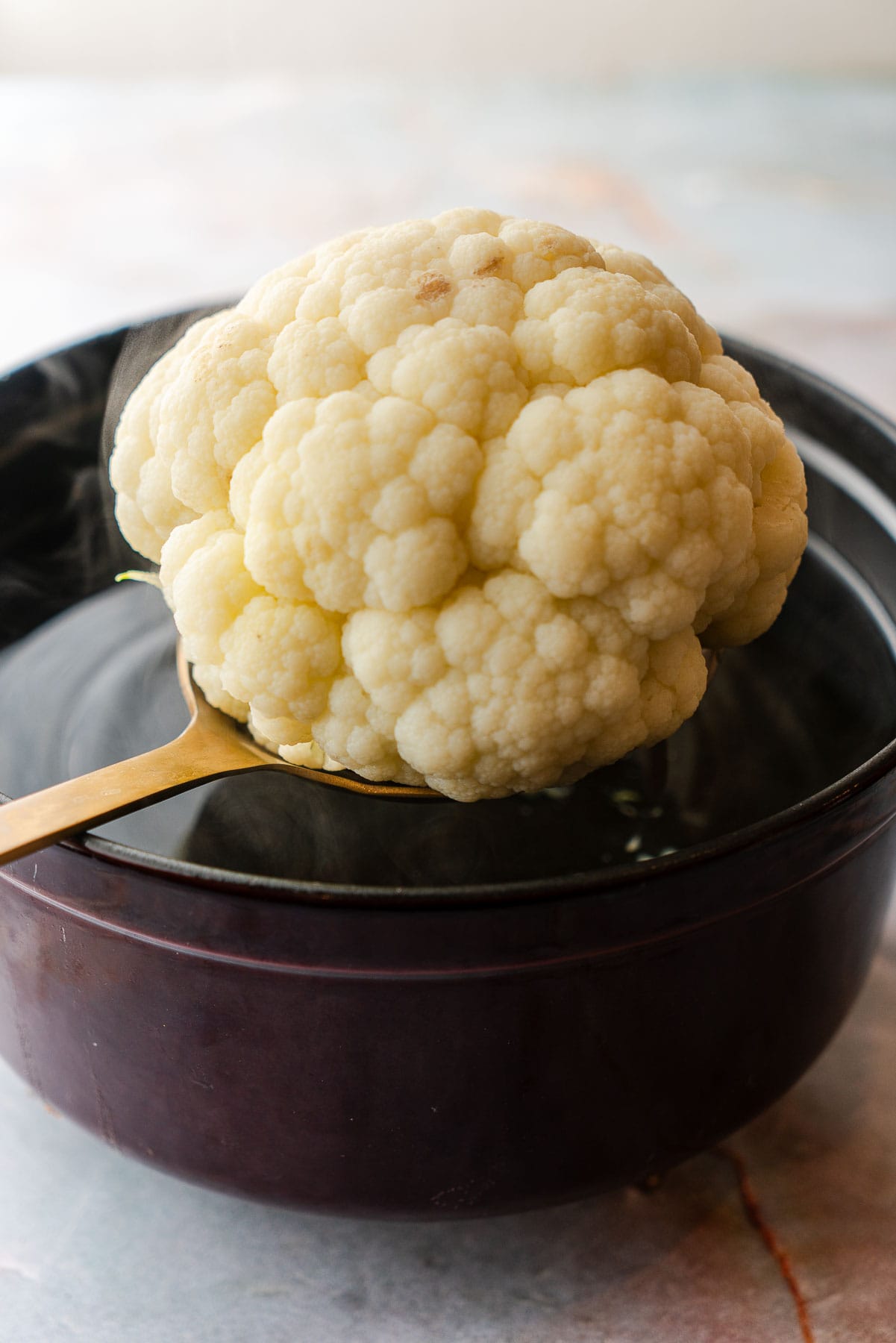 A slotted spoon lifting a steaming head of cauliflower out of boiling water.