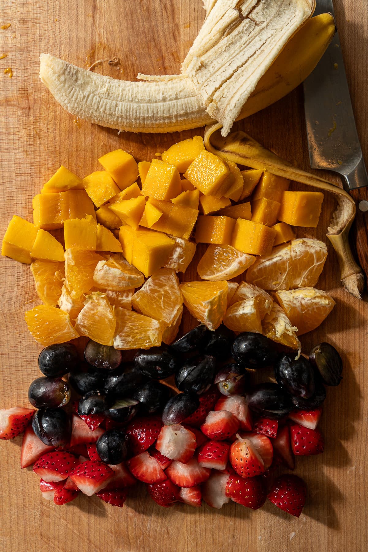 A cutting board with chopped assorted fruit, an uncut banana, and a knife.