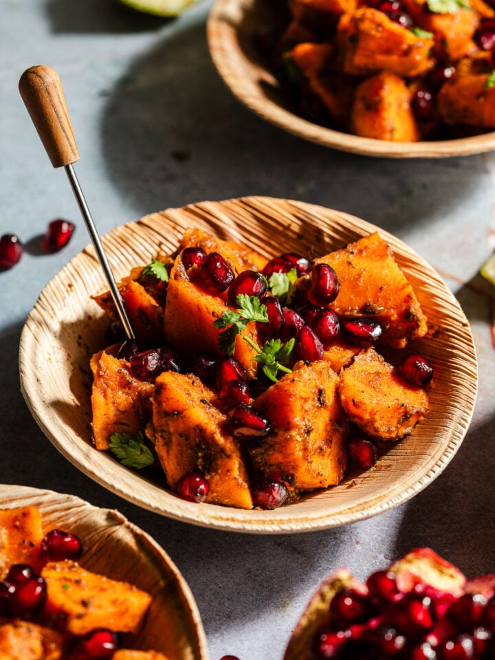 Sweet potato chaat with pomegranates in a palm leaf bowl with an appetizer pick.