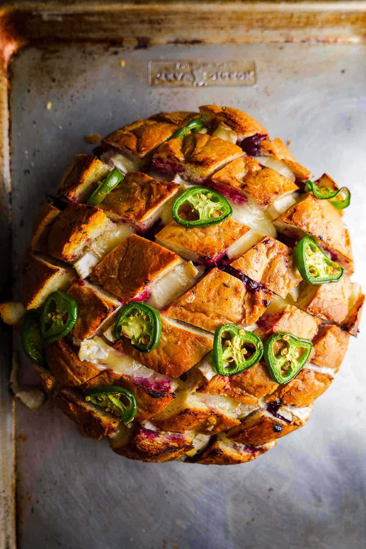 Baked loaf of bread with slices that are filled with cranberry sauce, brie, and topped with jalapeños.