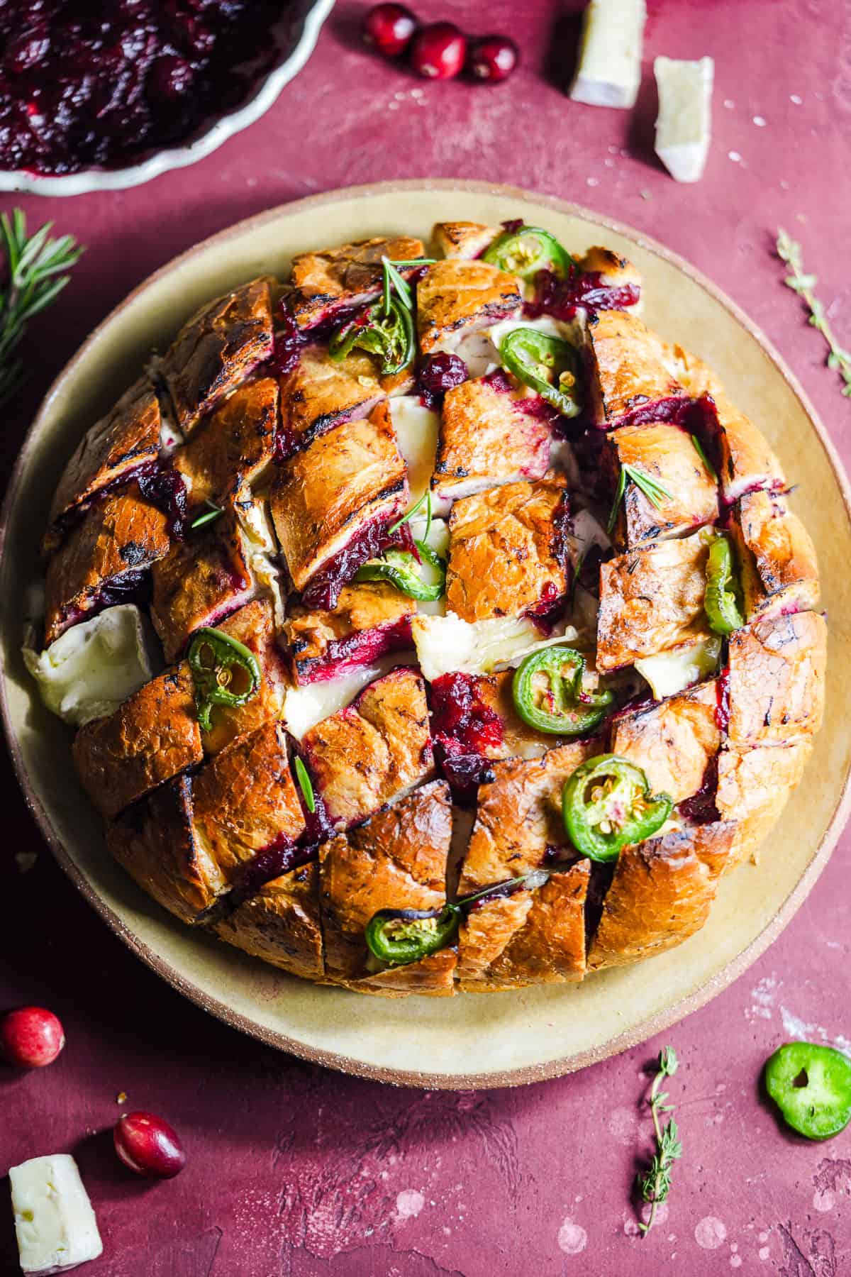 Baked loaf of bread with slices that are filled with cranberry sauce, brie, and topped with jalapeños.