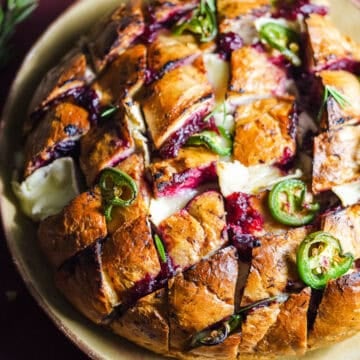 Baked loaf of bread with slices that are filled with cranberry sauce, brie, and topped with jalapeños.