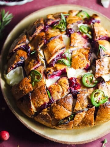 Baked loaf of bread with slices that are filled with cranberry sauce, brie, and topped with jalapeños.