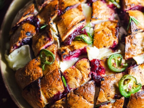 Baked loaf of bread with slices that are filled with cranberry sauce, brie, and topped with jalapeños.