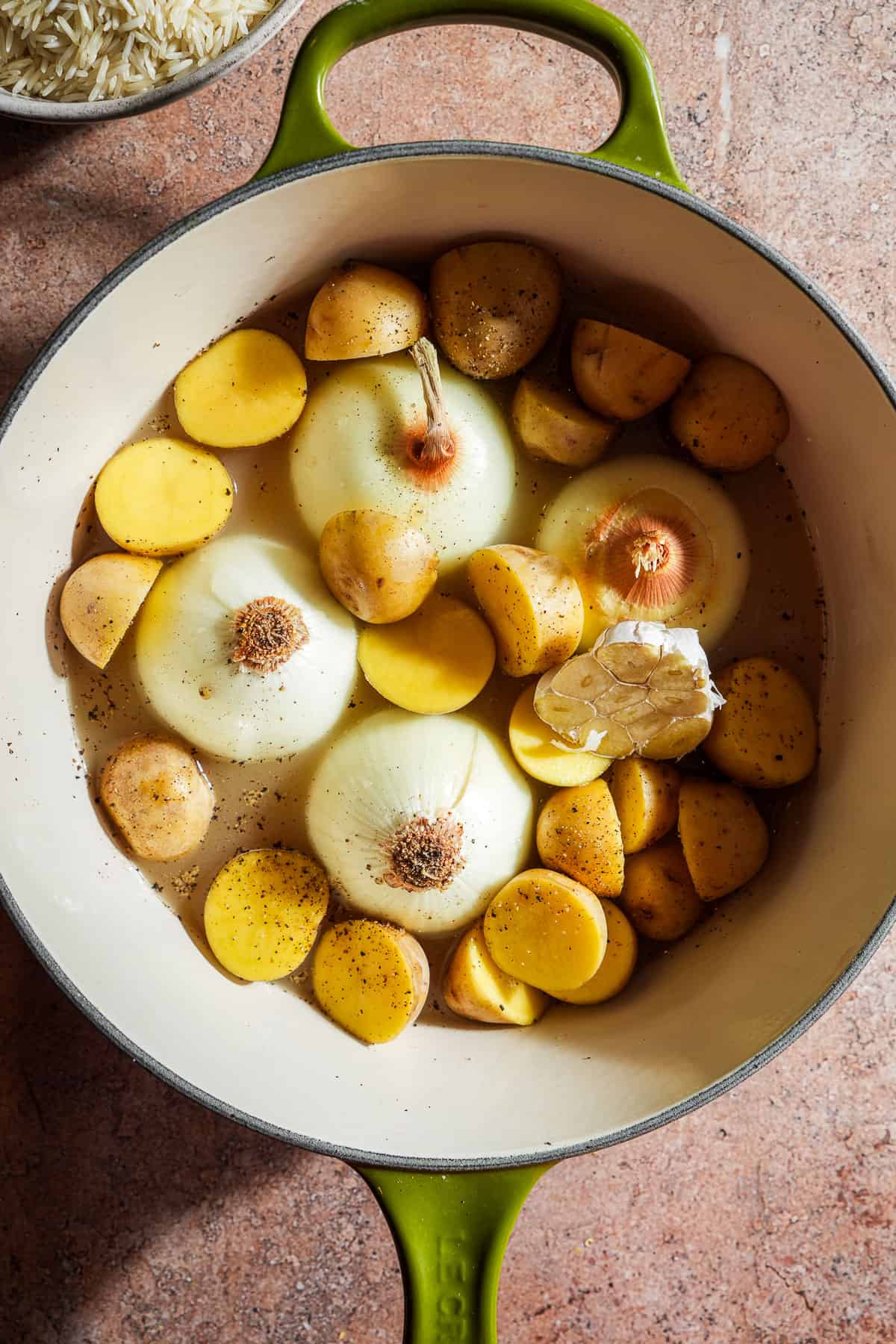 A dutch oven filled with halved onions, potatoes, garlic, and filled with broth and seasonings.