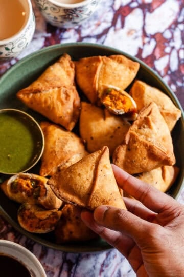 A hand grabbing a samosa from a platter with chutney.