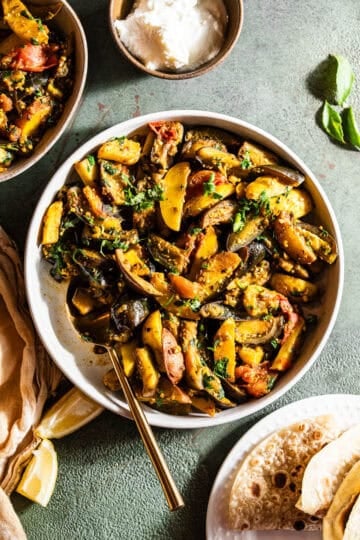 A bowl of food with potatoes, eggplant and tomatoes served with roti and dahi.
