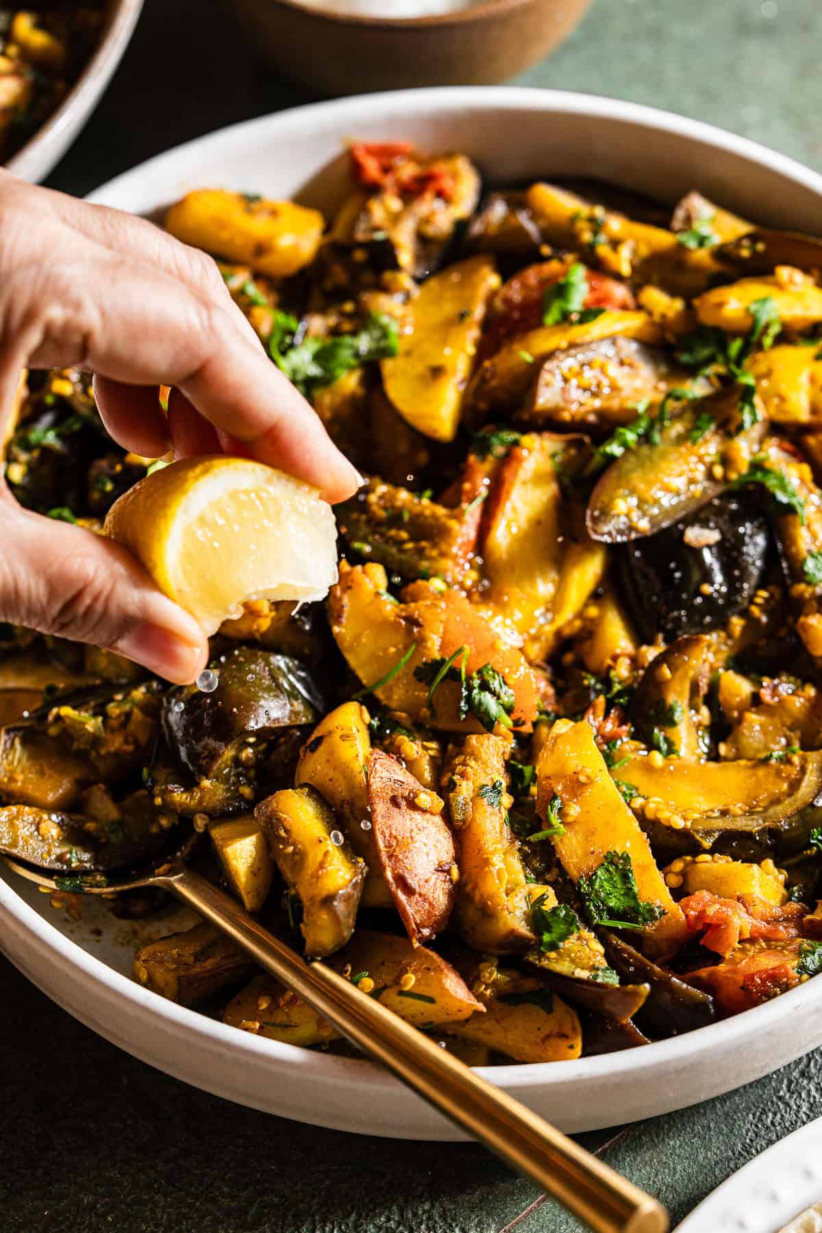A person is squeezing lemon juice over aloo baingan in a bowl.