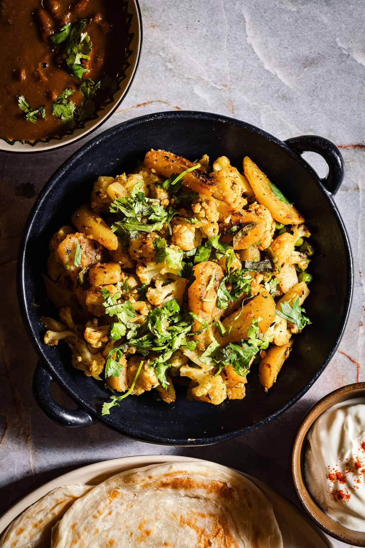 Aloo gobi with paratha, dahi, and rajma in separate bowls and plates.