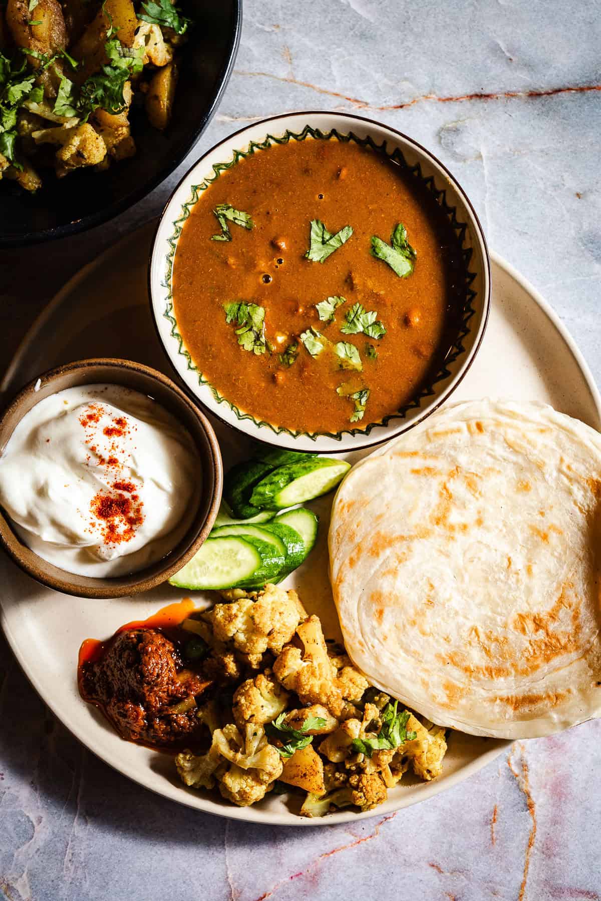 Aloo gobi with paratha, dahi, and rajma in a plate with cucumbers and achaar.