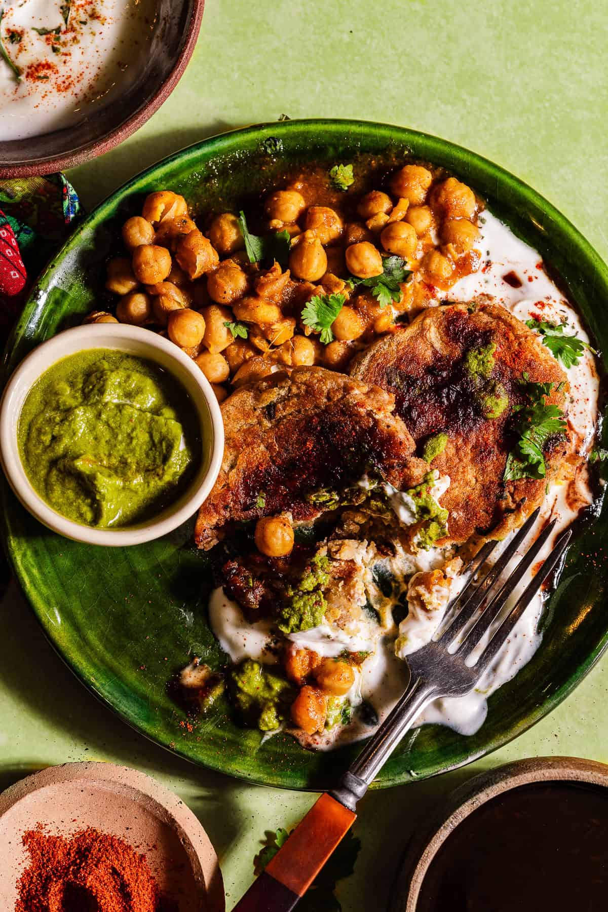 A fork digging into aloo tikkis with chutneys layers on dahi and chole.