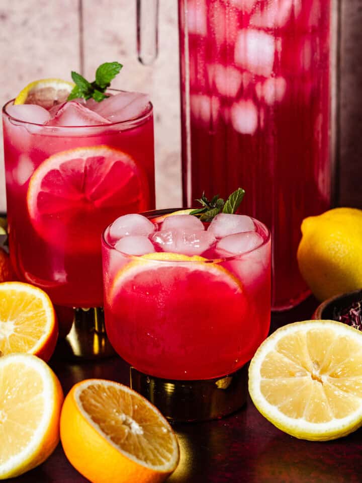 Hibiscus lemonade in glasses and a pitcher with ice surrounded by citrus.
