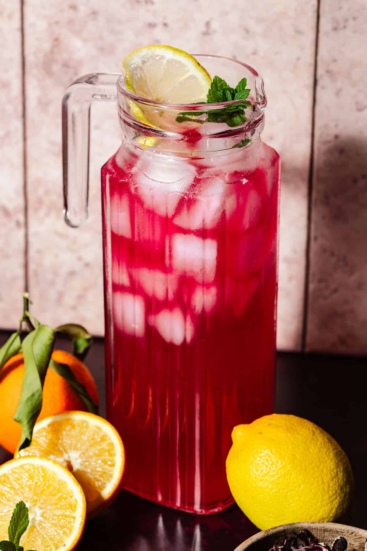 Hibiscus lemonade in a pitcher with ice surrounded by citrus.