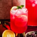 Hibiscus lemonade in glasses and a pitcher with ice surrounded by citrus.
