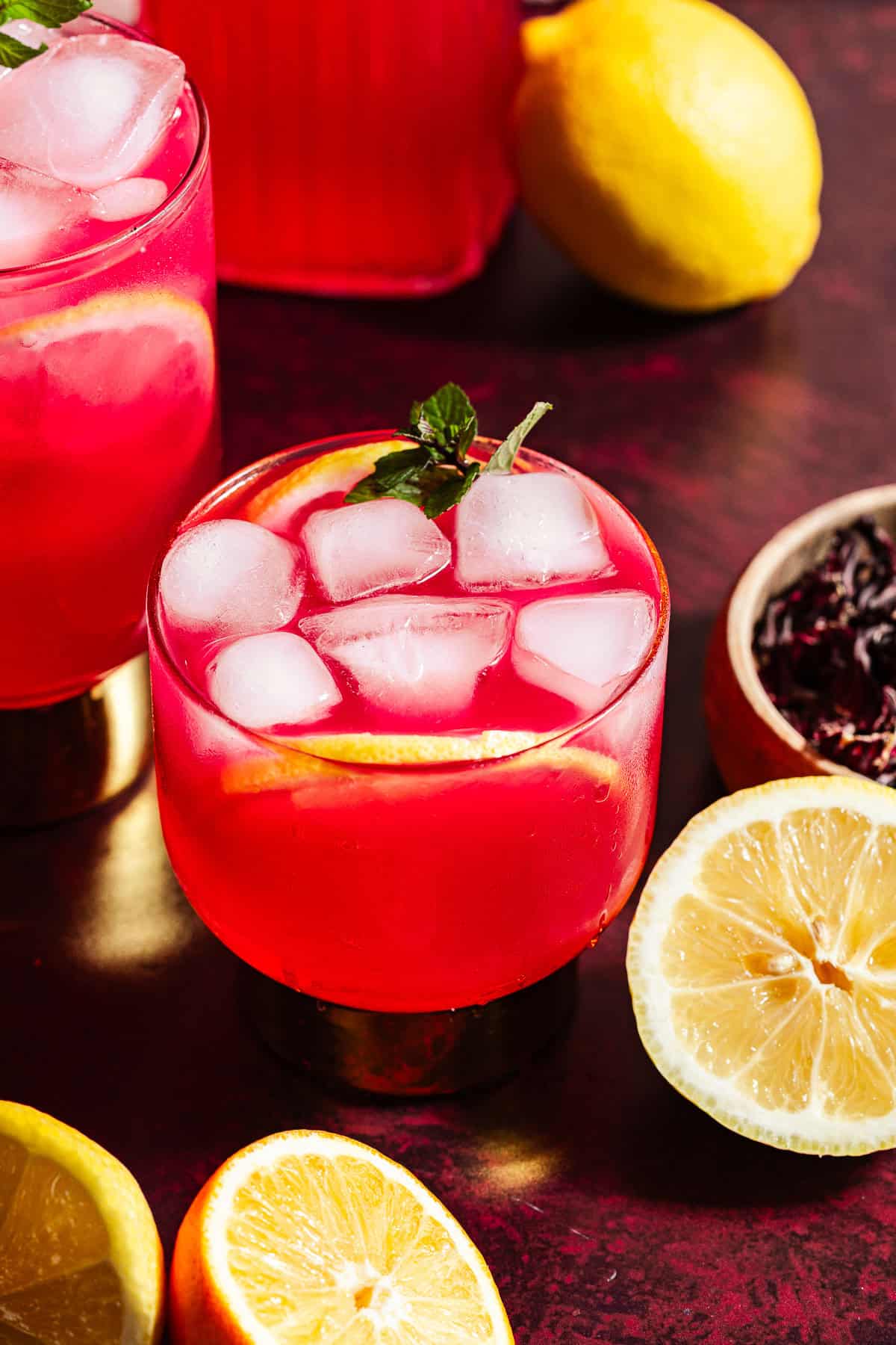 Hibiscus lemonade in glasses and a pitcher with ice surrounded by citrus.