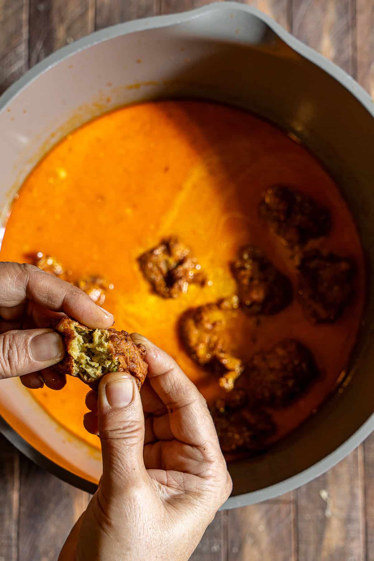 A hand slightly breaking open a kofta to add it to a pot with tomato gravy and koftas.