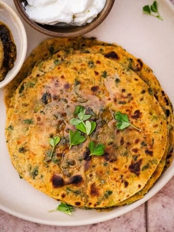 A stack of methi paratha with ghee, fresh methi, and served with dahi and achaar.