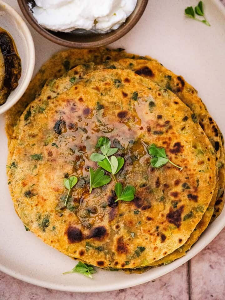 A stack of methi paratha with ghee, fresh methi, and served with dahi and achaar.